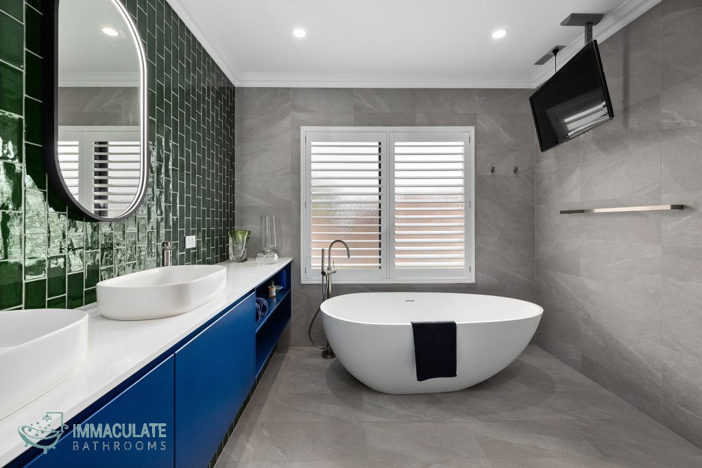 Modern bathroom renovation with freestanding bathtub, green feature tiles, blue vanity, and wall-mounted TV.