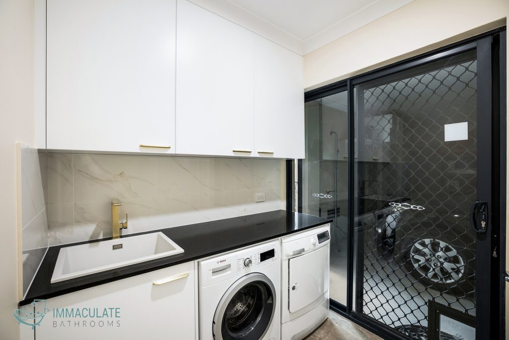 Modern laundry renovation featuring white cabinetry, marble-look splashback, black stone benchtop, gold tapware, and built-in washer and dryer beside sliding mesh door.