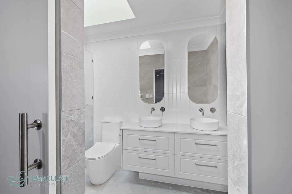 Modern Perth bathroom renovation featuring dual stone basins, white vanity cabinetry, vertical white wall tiles, grey floor tiles and twin arched mirrors beneath a skylight.
