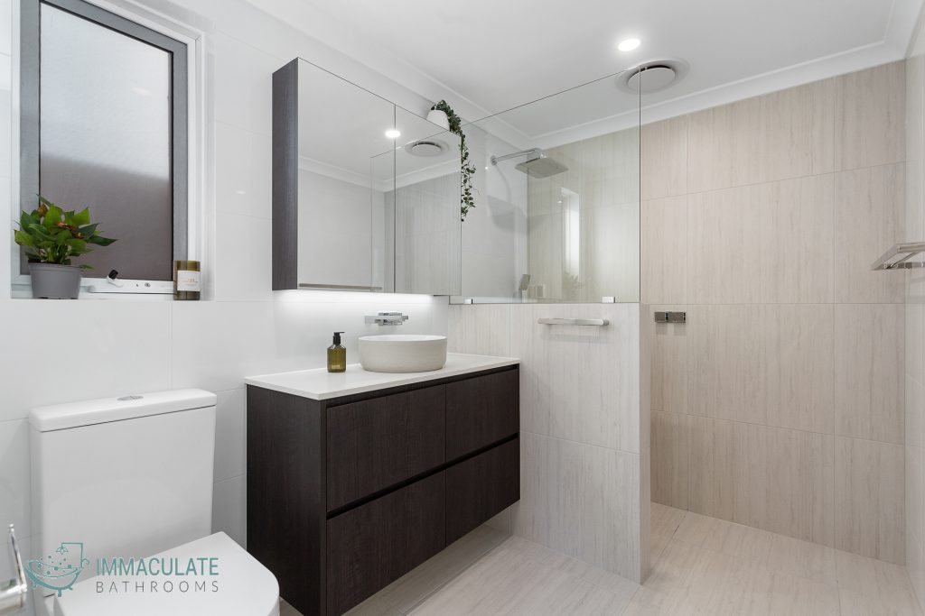 Modern bathroom renovation featuring a walk-in shower with glass screen, wall-hung dark timber vanity, round basin, and light stone wall tiles — Immaculate Bathrooms Perth.