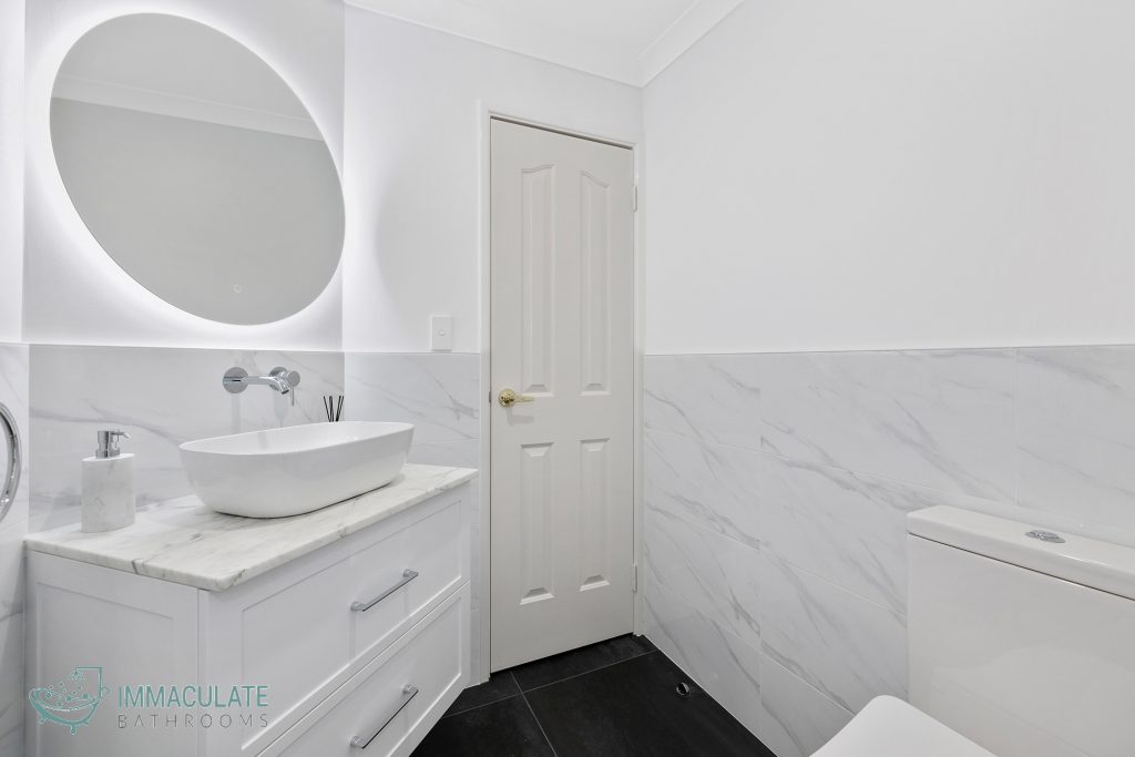 Modern white bathroom with illuminated round mirror, marble vanity top, and wall-mounted basin by Immaculate Bathrooms Perth