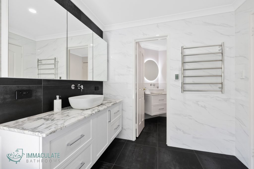 Modern bathroom renovation featuring a marble-look vanity top, white shaker cabinetry, vessel sink, large mirrored cabinet, black feature tile splashback, heated towel rail, and view into matching ensuite.