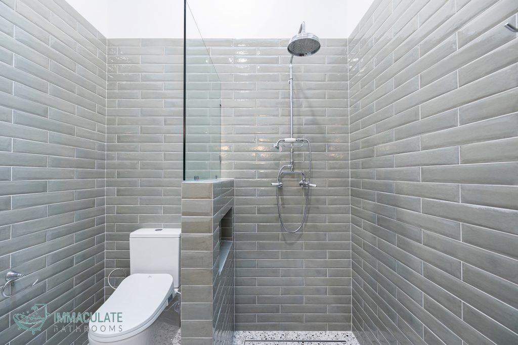 Modern bathroom with light grey subway tiles, walk-in shower with rainfall showerhead, half-height tiled partition with niche, and white toilet.
