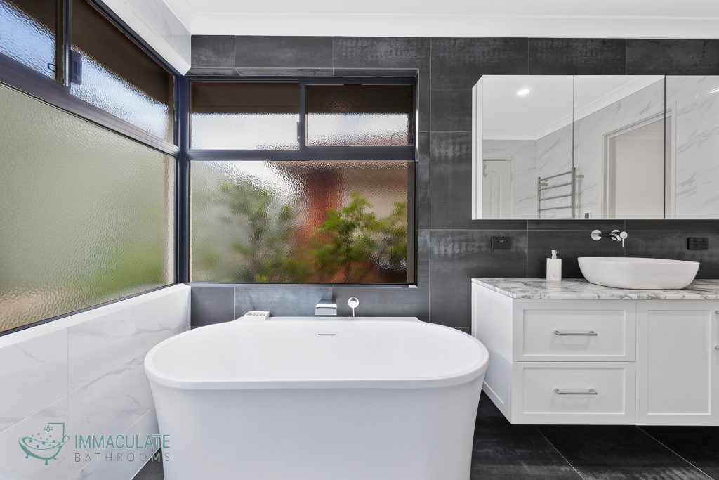 Modern bathroom with freestanding white bathtub, large frosted windows, dark feature tiles, and white vanity with marble benchtop and round basin.