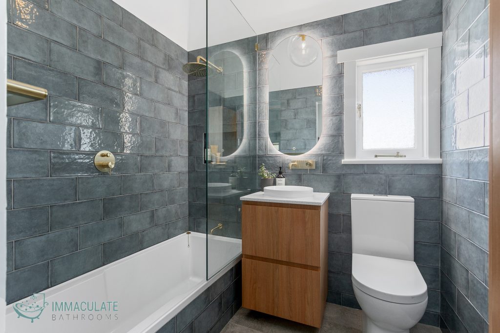 Modern grey-tiled bathroom with shower-bath combo, timber vanity, and gold fixtures.