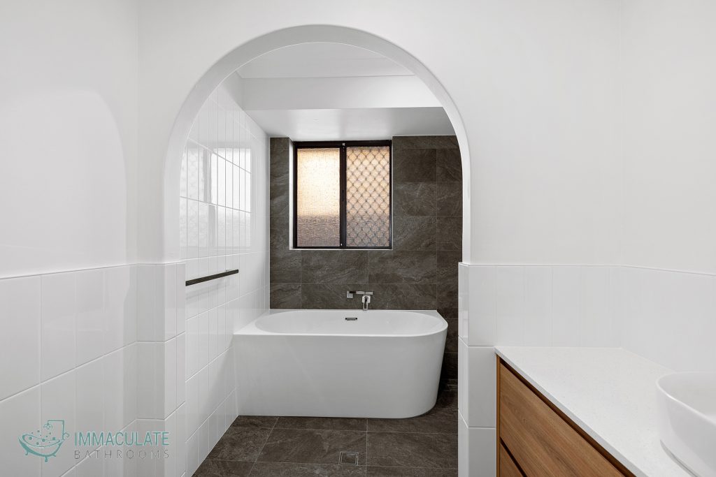 Modern bathroom featuring an arched entry with a freestanding bathtub, white wall tiles, stone-look floor and wall tiles, and a timber vanity by Immaculate Bathrooms Perth.