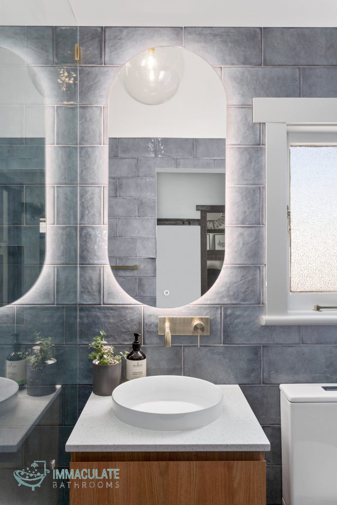 Modern bathroom renovation featuring a curved backlit mirror, textured grey subway tiles, brass wall-mounted tapware, and a white round vessel basin on a timber vanity.