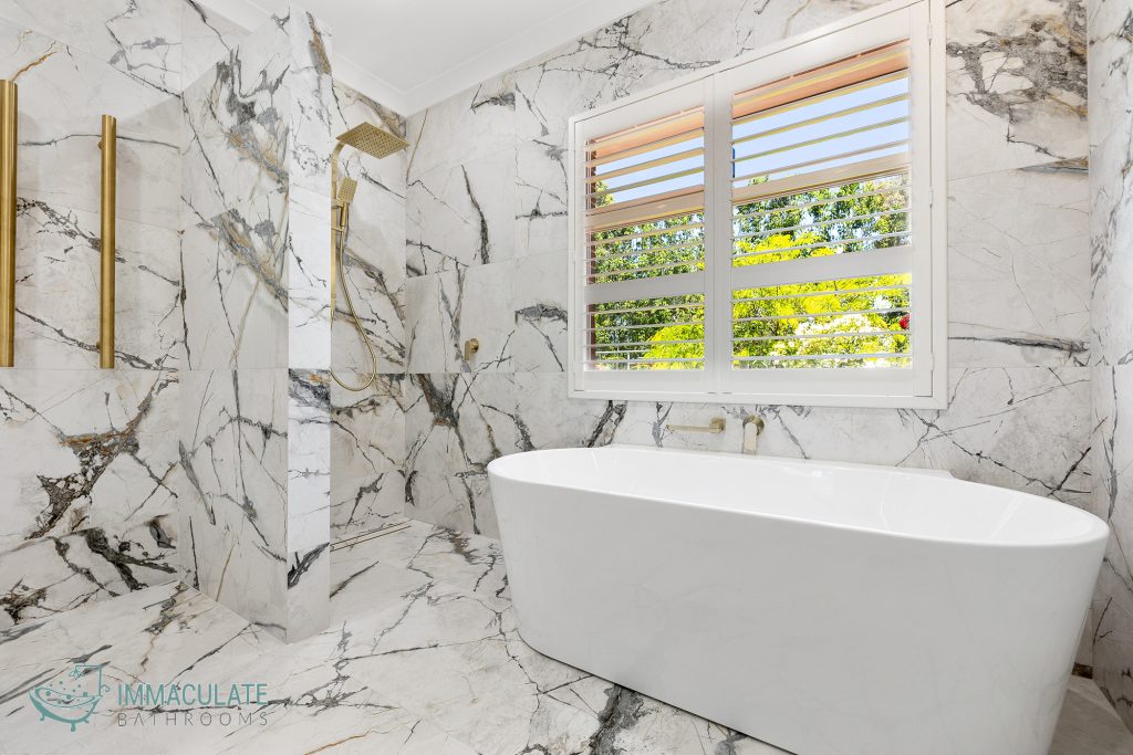 Luxury marble-look bathroom with freestanding bath, gold tapware and large window by Immaculate Bathrooms Perth