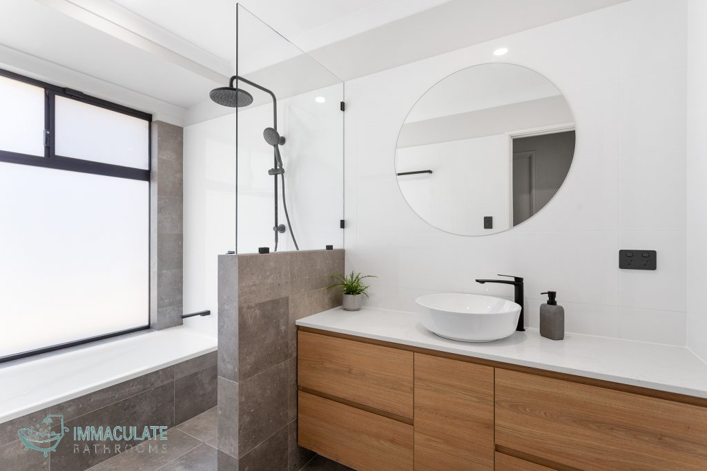 Modern bathroom with timber vanity, round mirror, matte black tapware and shower-over-bath by Immaculate Bathrooms Perth