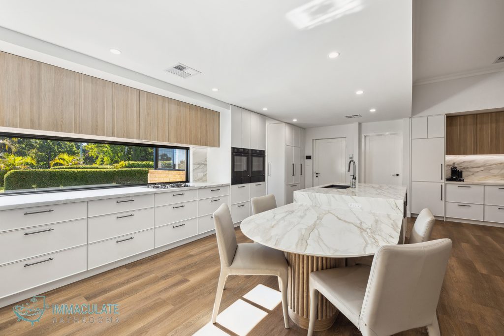 Modern white kitchen with large marble island bench, wood-grain overhead cabinets, integrated appliances, and open window splashback.
